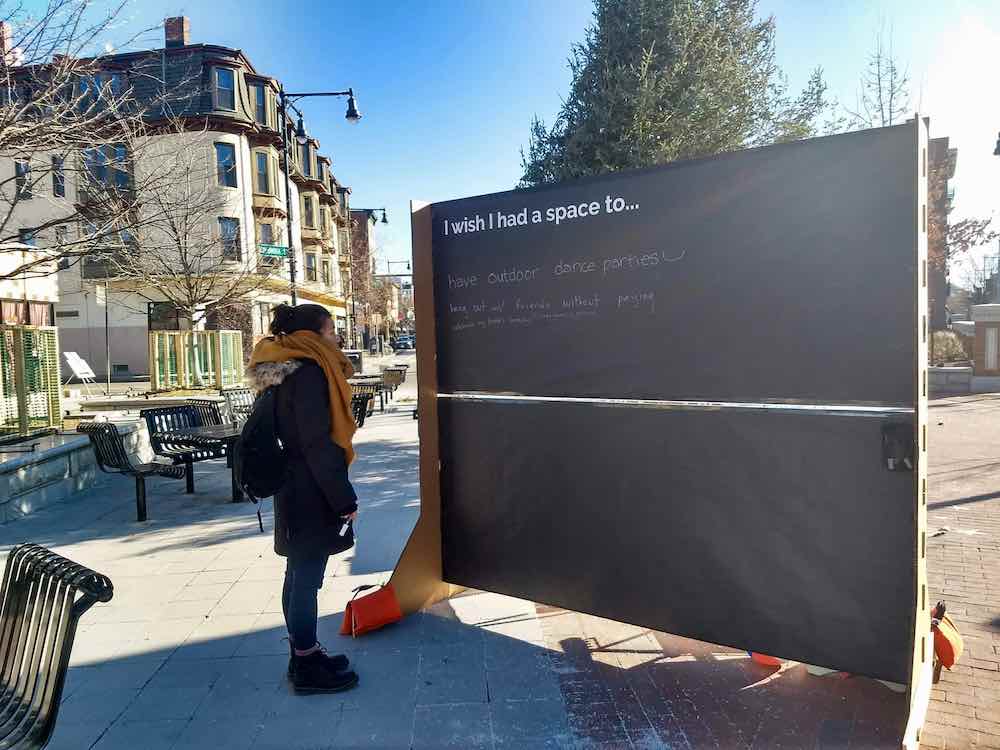 A woman looks at a large chalk board that reads "I with I had a place to..." in Lafayette Plaza in Cambridge