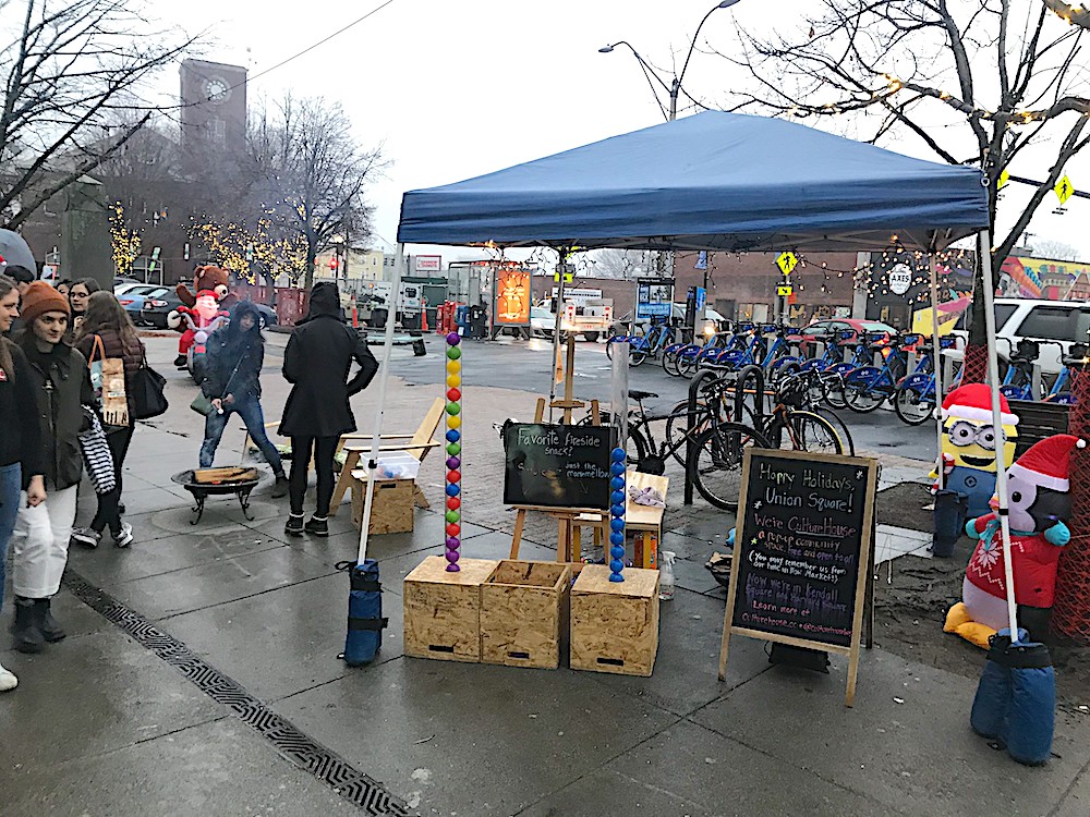 A fire pit, chairs, and tubes with balls in them for voting under a tent at the Union Square Holiday Stroll