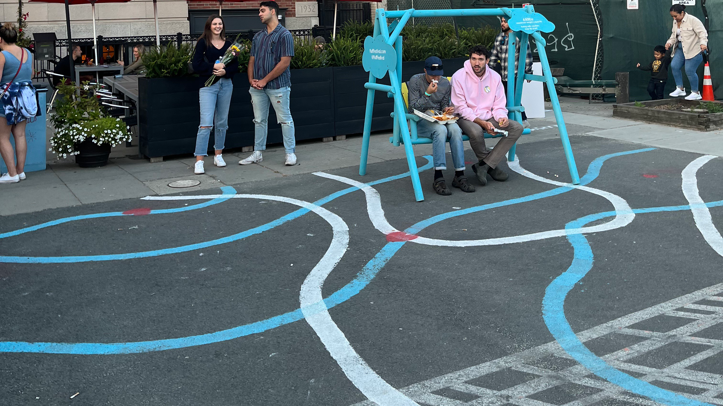 a plaza with white and blue painted lines on the ground. Two people sit on a blue swing.