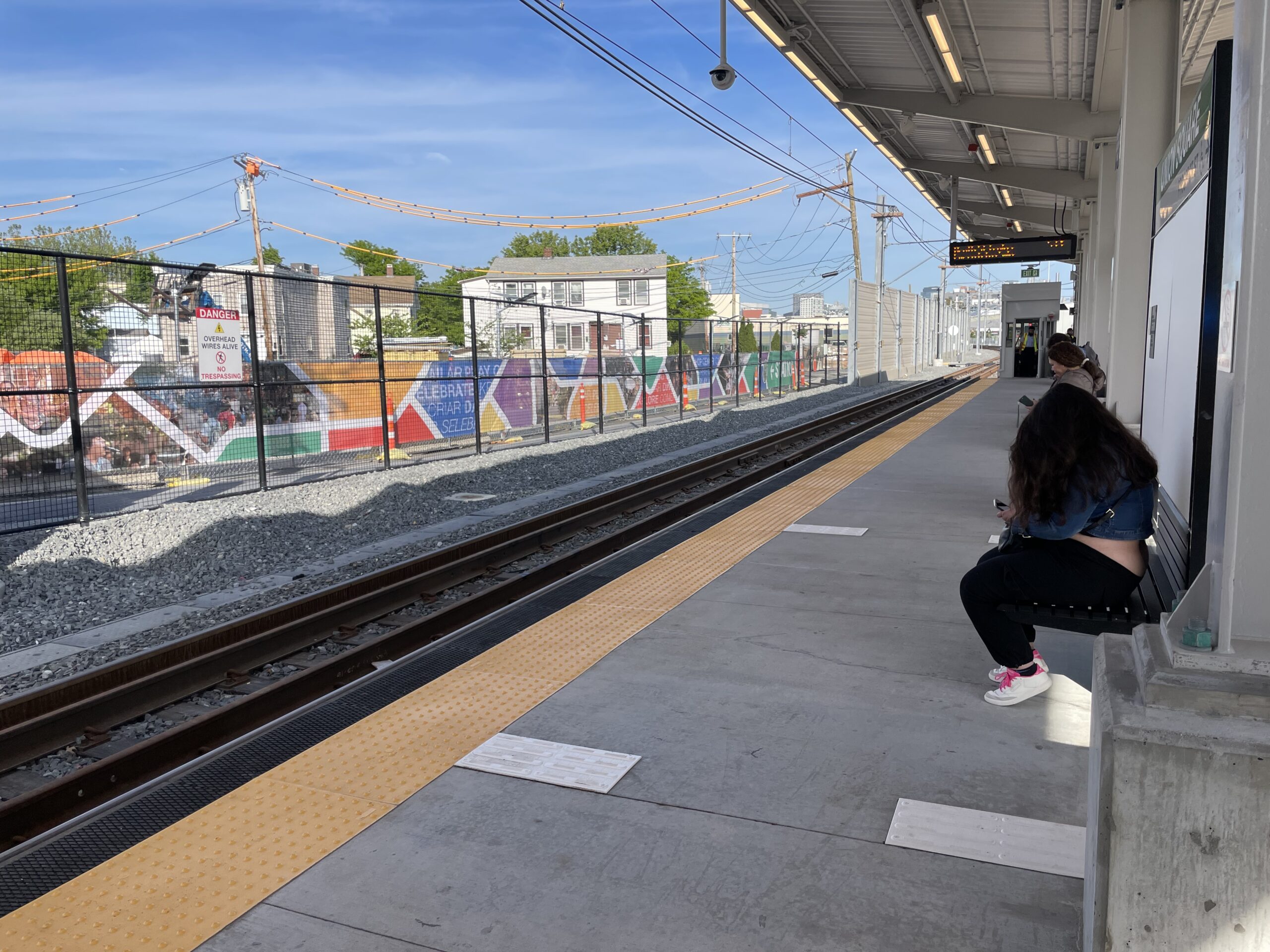 People sit on the train platform with the scrim visible on the other side of the tracks.