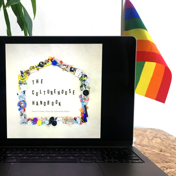 a gray laptop on a wood grain table. A rainbow flag and small plant sits behind the laptop. The laptop has an image of a book cover that reads "The CultureHouse Handbook."