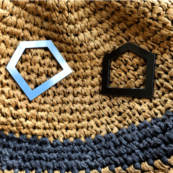 Two CultureHouse black enamel pins against a brown straw texture. The enamel pins are in the shape of a black five point house.