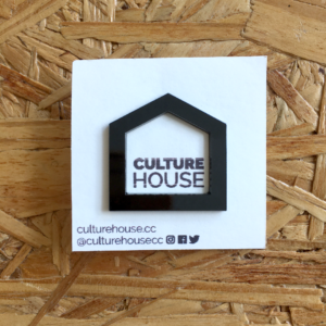 a black enamel pin against a white cardstock paper. The paper reads "CultureHouse." The pin and paper are against a woodgrain table.