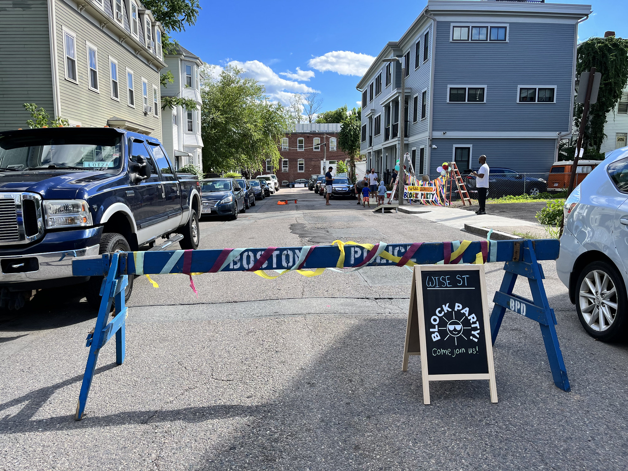 A decorated street divider sits in front of a block party alongside an a-frame sign that reads Wise St. Block Party Come join us!