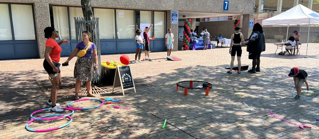 People standing in an open plaza with Game kit supplies scattered around while people play cornhole and chat.