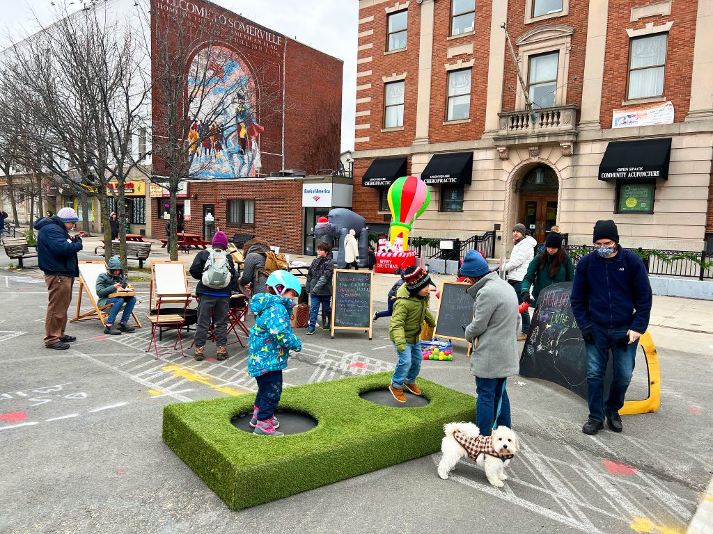 Two kids jump on a trampoline while adults mill about a fire pit and answer prompts about winter. In the background, there are large holiday inflatables.