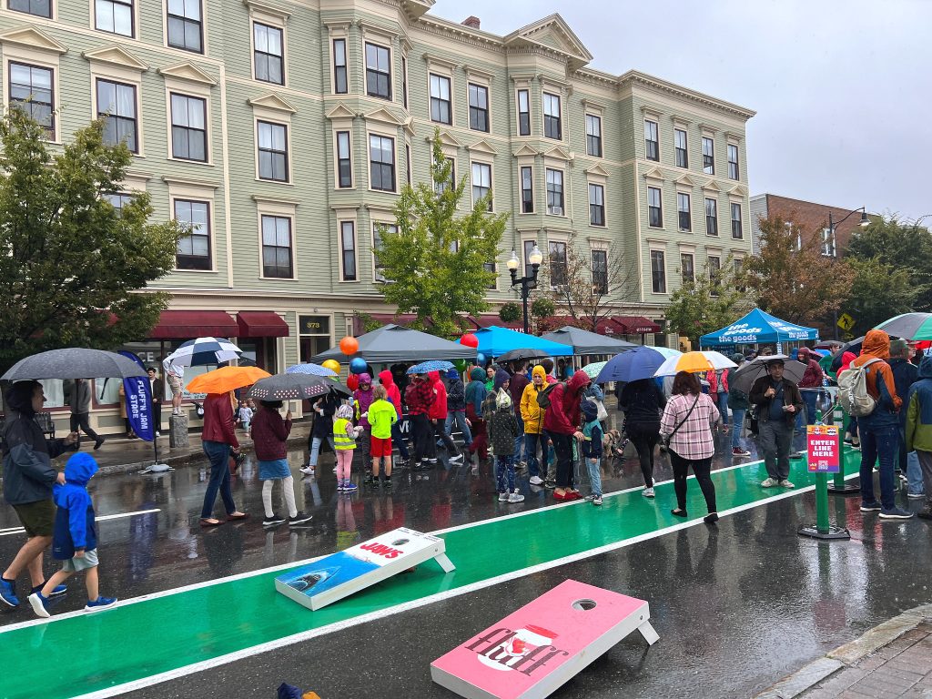 A crowd of people holding umbrellas move down Somerville Ave. Cornhole sets are on the street and vendors along the sides of the street