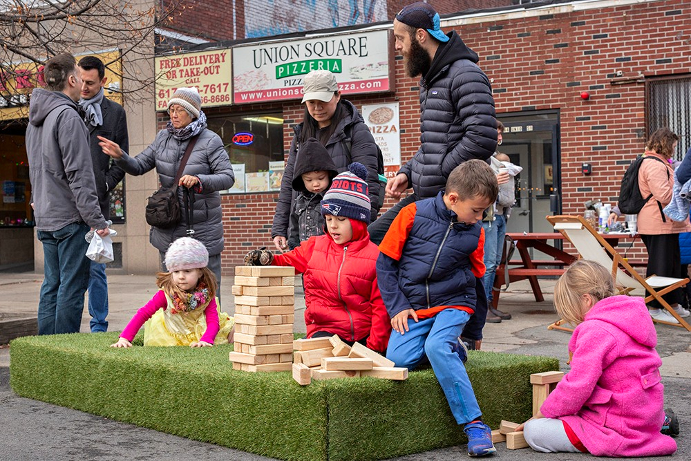 Children play jenga on a trampoline while parents look on in front of Union Square Pizza.