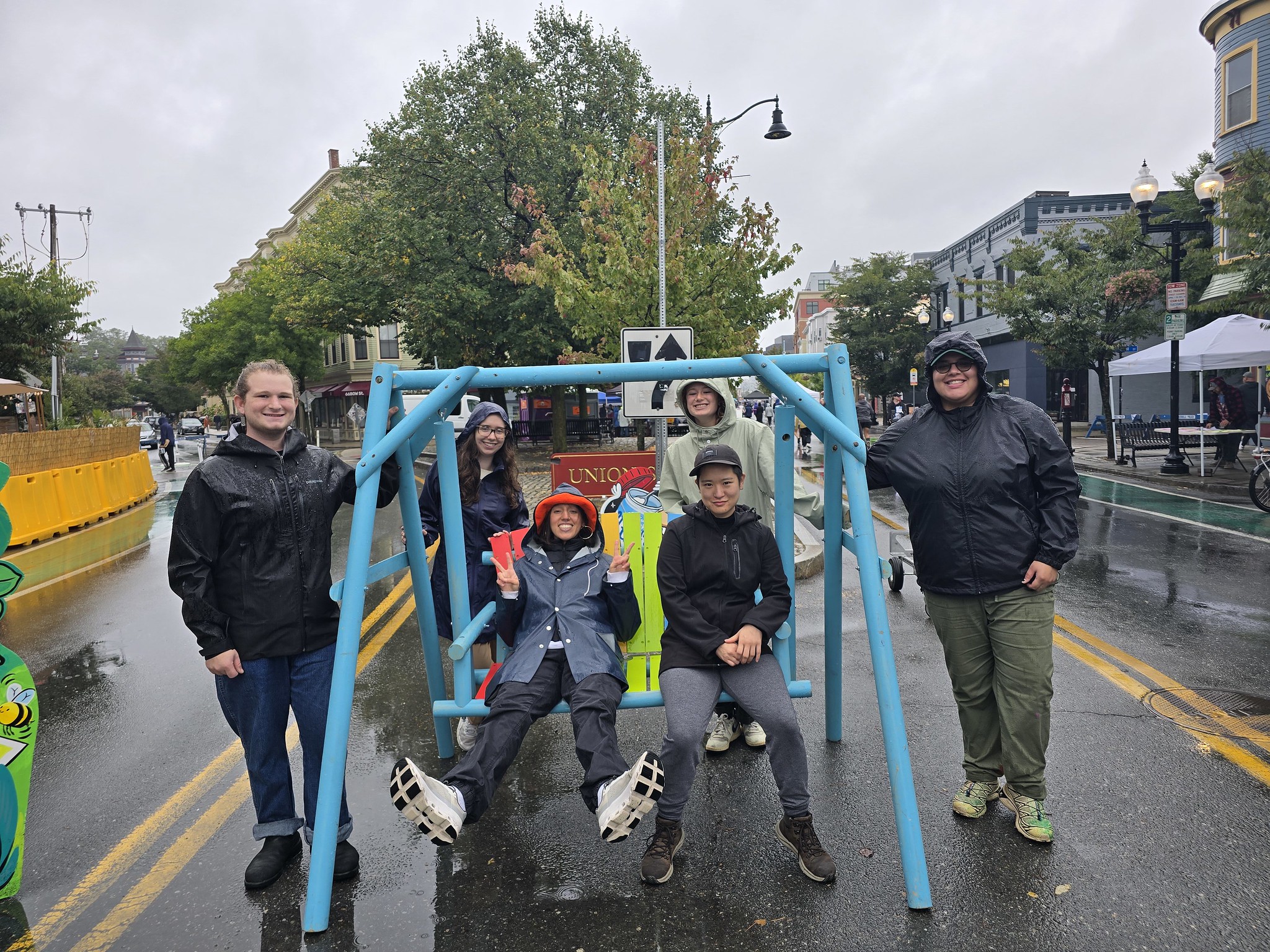 Two people sit on a swing while four people stand around in the rain smiling.