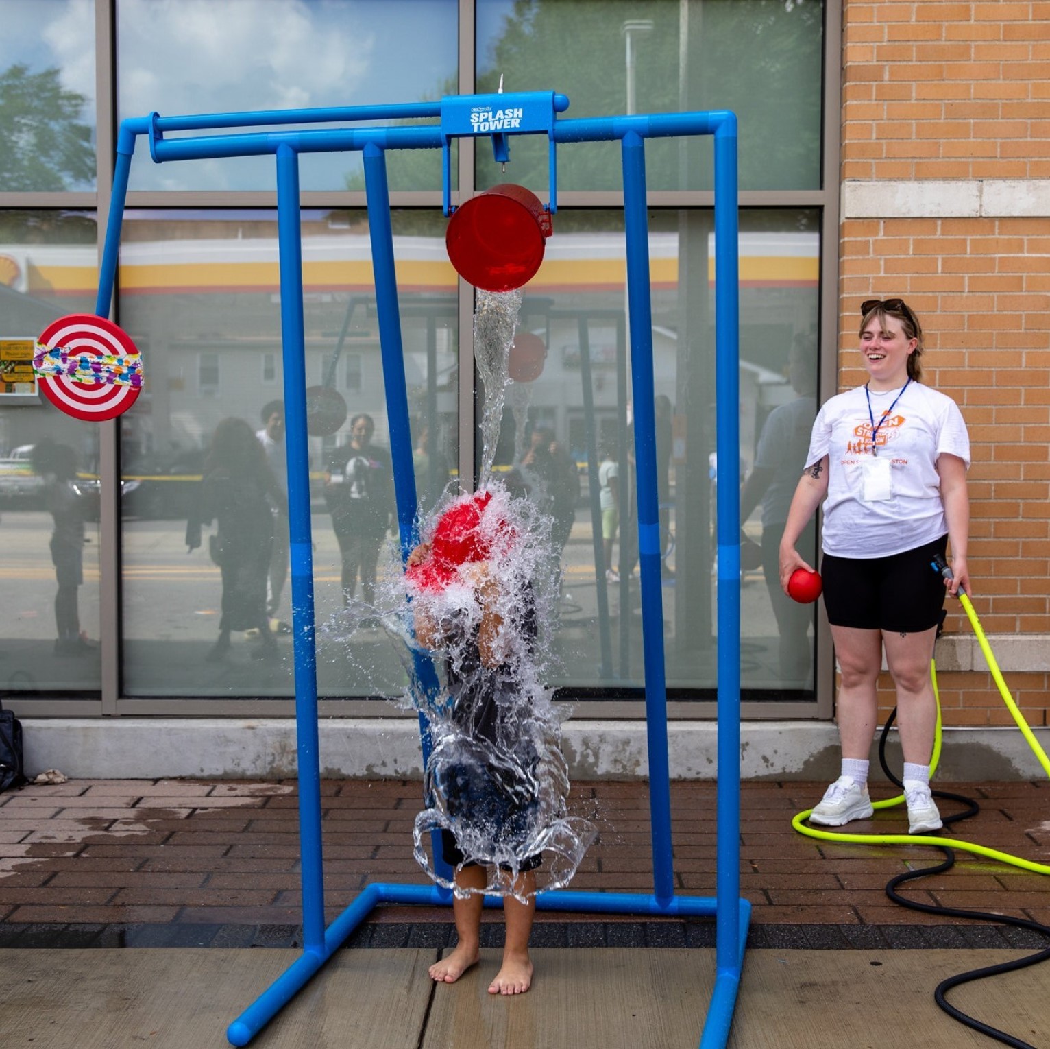 An Open Streets Volunteer stands smiling behind a child under the Splash Tower getting a bucket of water dumped on their head.