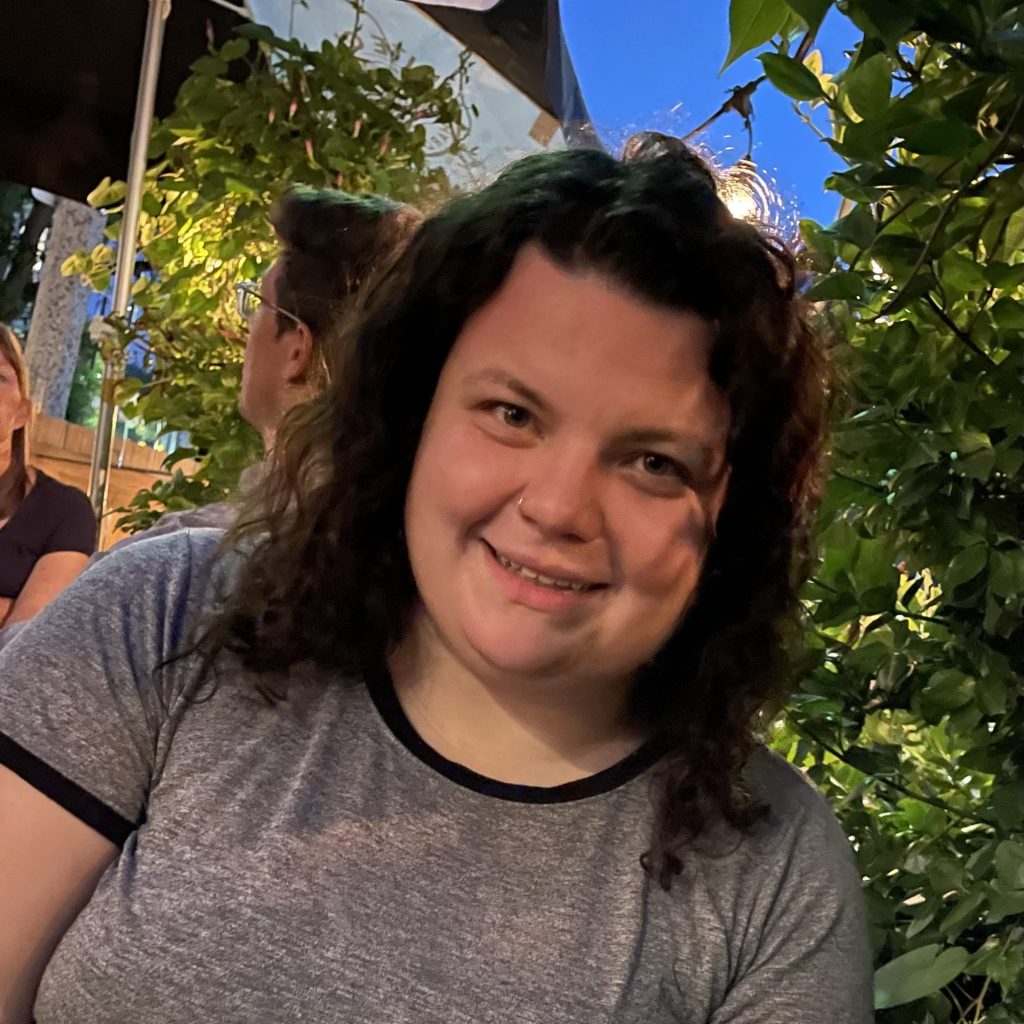 A brunette, white female-presenting person sitting in an outdoor cafe during the early evening. They are smiling at the camera.