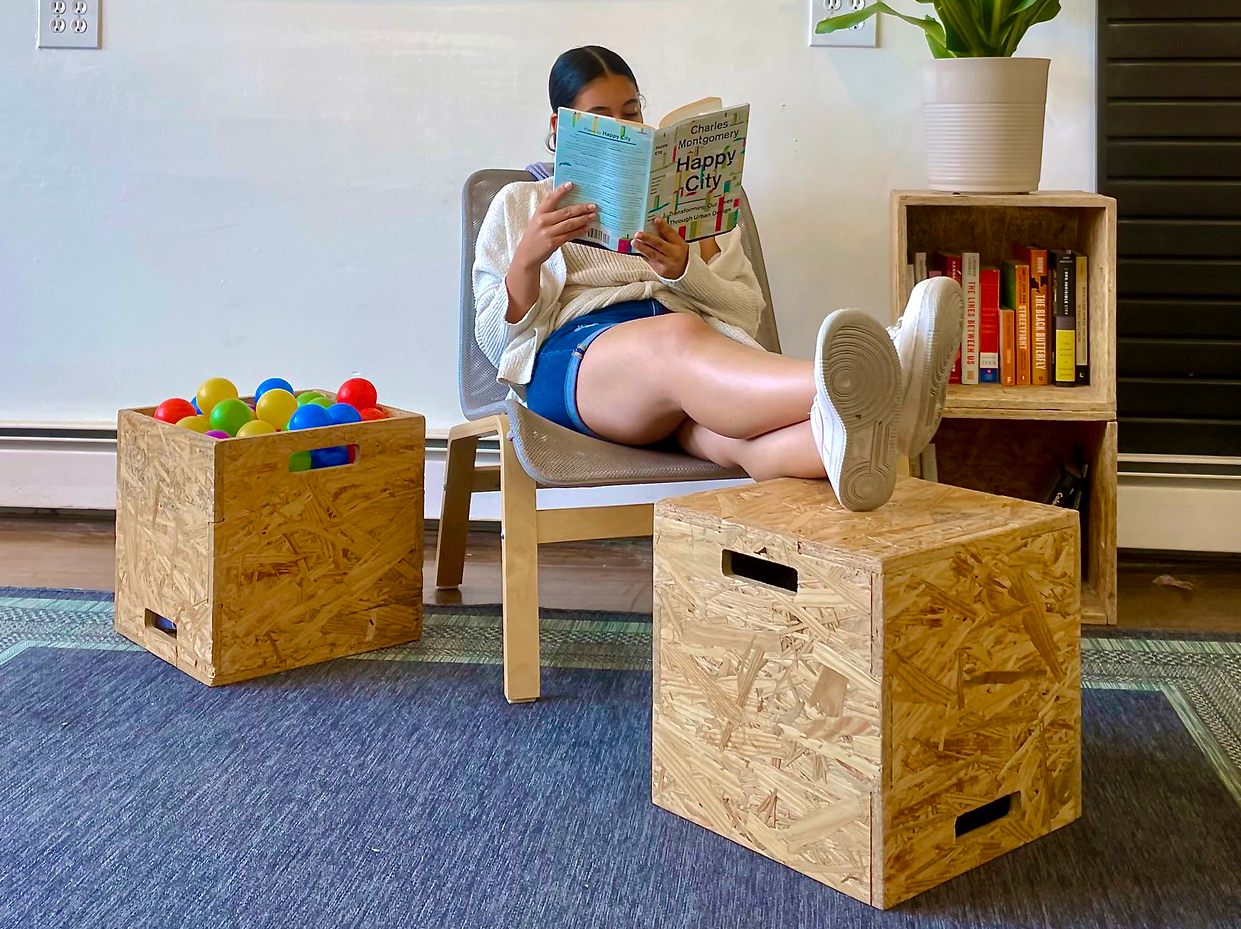 A woman sits on a chair with her feet up on a cube stool reading a book. Next to her, a stool is used to store colorful balls and on the other side another is used as a bookcase.