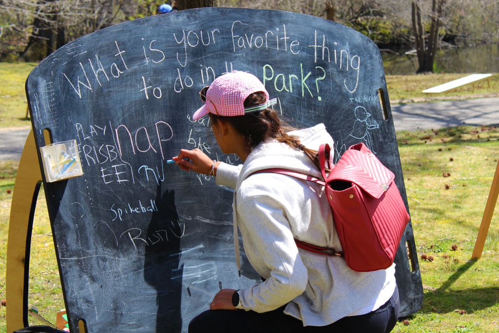 A woman crouches while writing on a chalkboard in a park.