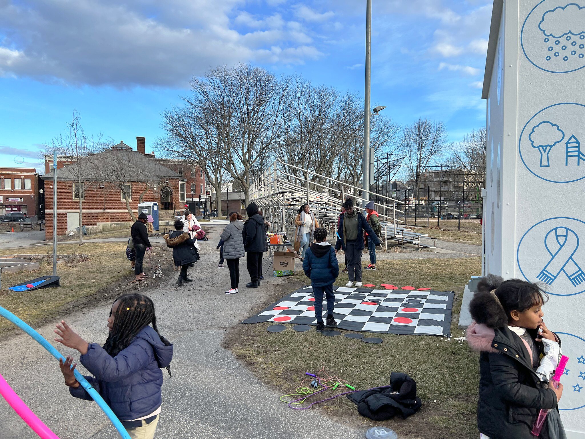 People and children standing around playing games and eating snacks. Giant checkers on the ground next to the new play shed.