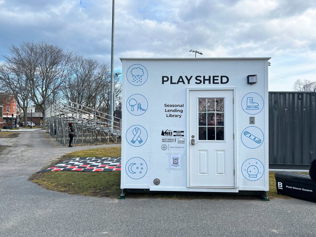 The outside of a small shed reads "Play Shed: Seasonal Lending Library" lists partners. Winter icons decorate the outside of the shed and a giant checkerboard is in the background.