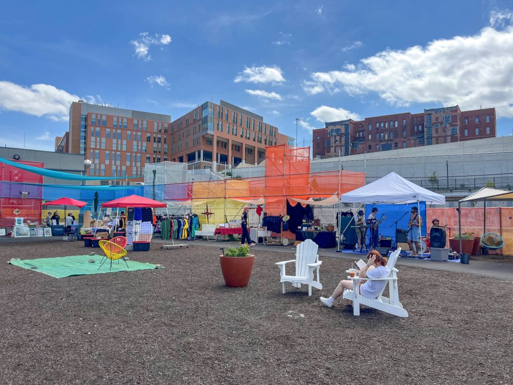 Person reading a book on a white chair surrounded by a market under colorful scaffolding and a band