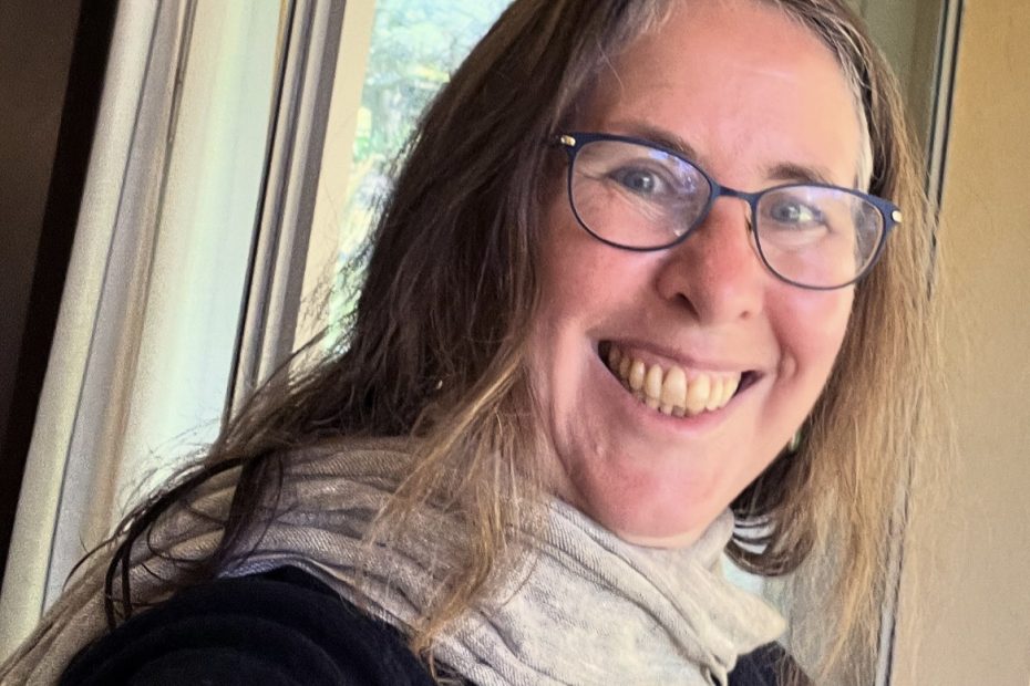 Face of a woman smiling, wearing glasses, a black sweater, and grey scarf