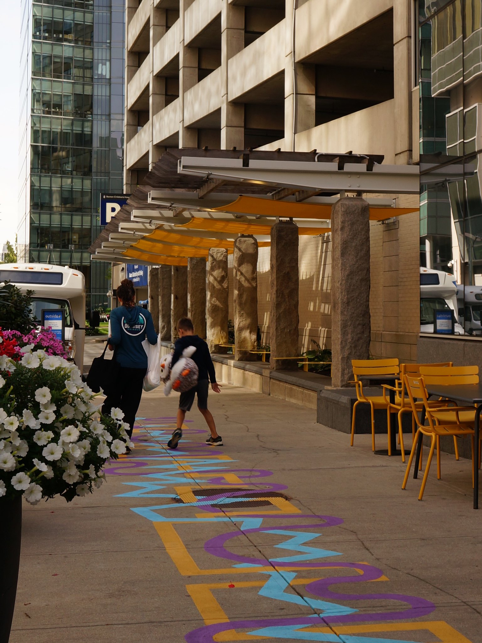 A mother and child walk on a colorful ground mural with bright sun shades and tables in the background.