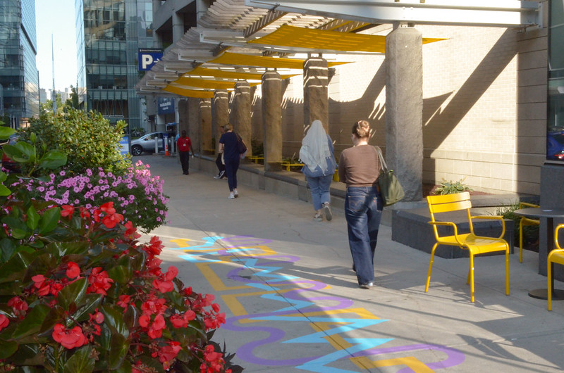 People on a walkway with flowers in the foreground, bright yellow sun shades, and a colorful mural on the ground.