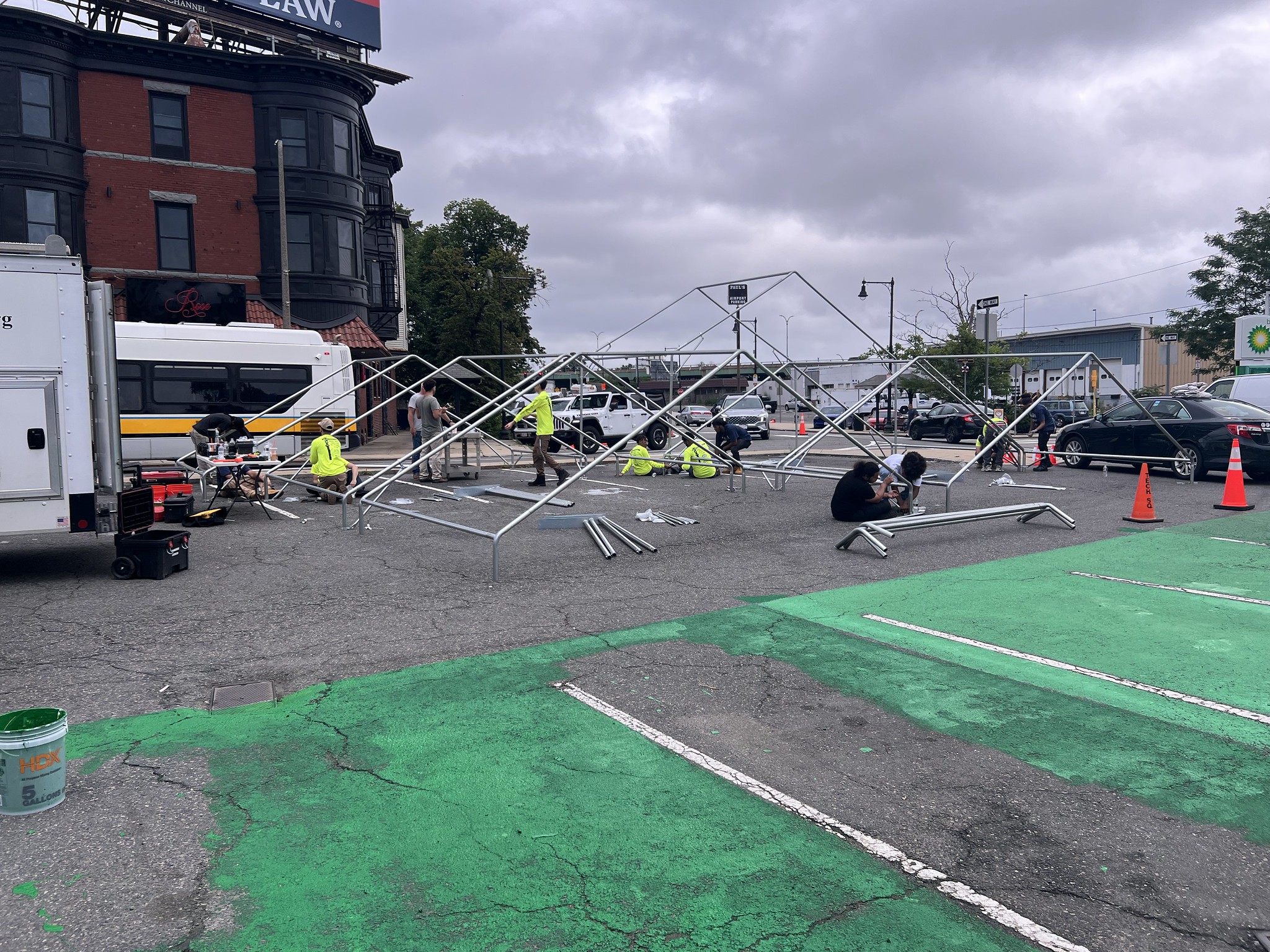 In the middle of a parking lot, people construct metal structures while a green mural is being put down.