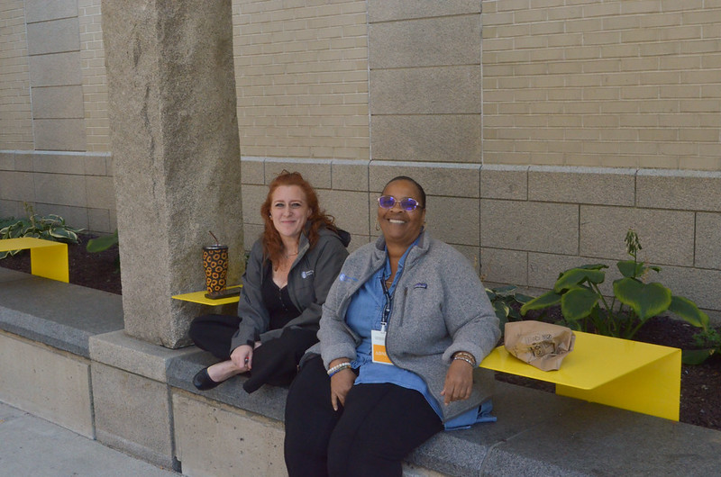 Two women sit next to each other on a concrete bench smiling with their lunches