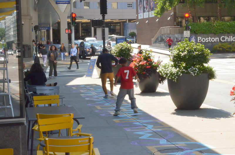 A boy and and a man jump and play on a colorful ground mural on a sidewalk next to tables and planters