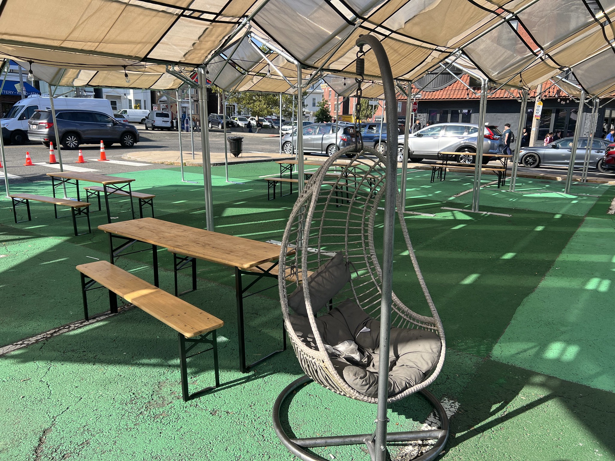 A hammock chair sits next to picnic tables underneath a tan shade structure.