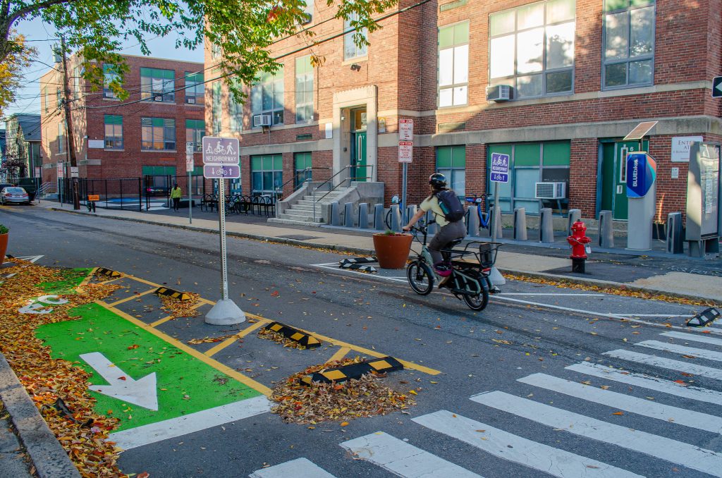 A person on a cargo bike entering Otis Street, passing a narrowed gateway with signs and a contraflow bike exit.