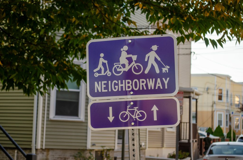 A sign with symbols of a kid on a scooter, a femme-presenting person riding a bike, and a person in a hat walking a dog, all above the word "NEIGHBORWAY."