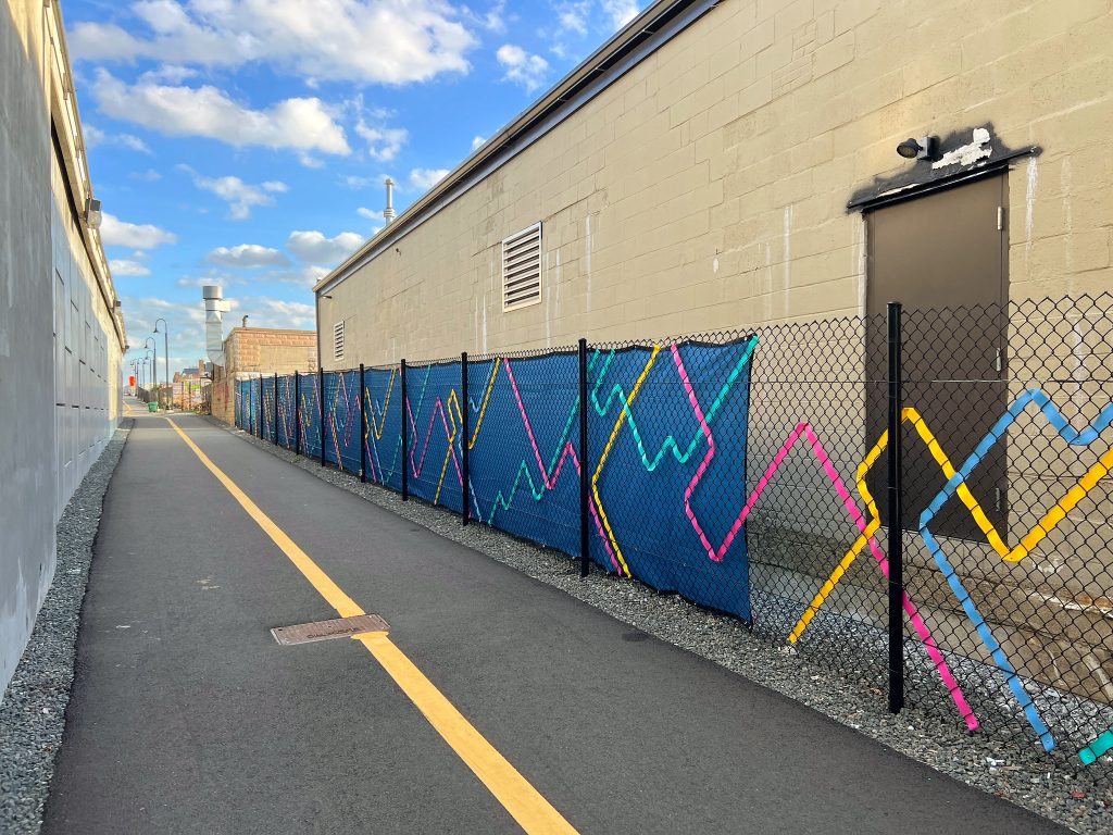 A shared path leads into the distance, a colorful fence weave is on the chain link fence next to the path