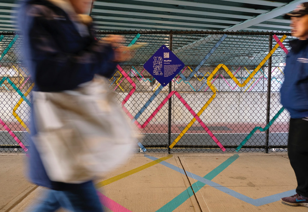 People walk by in front of a colorful fence and on a ground mural