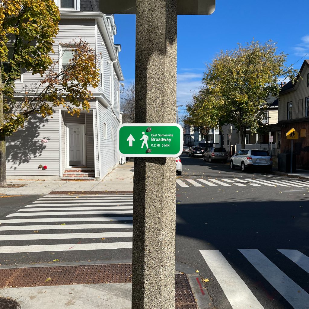 A sign posted on a pole reads "East Somerville Broadway 0.2 MI 5 MIN"