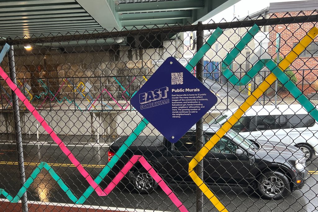 A sign describes the history of East Somerville public art and murals mounted on a chain link fence