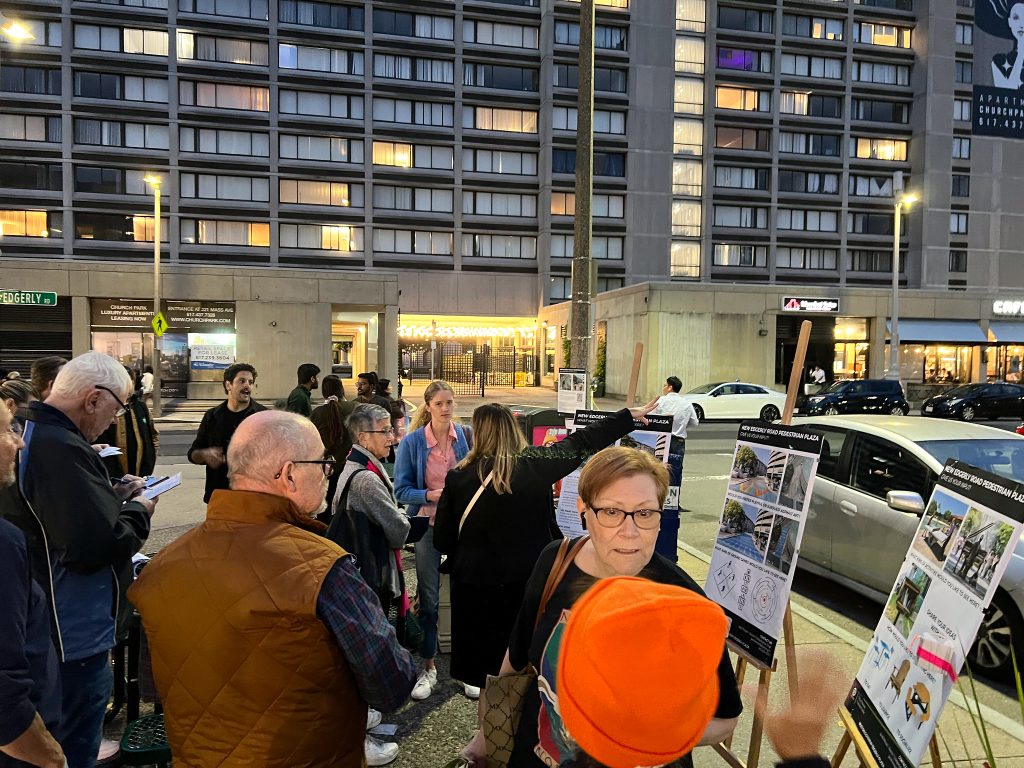 People gather around information boards during a community meeting outside