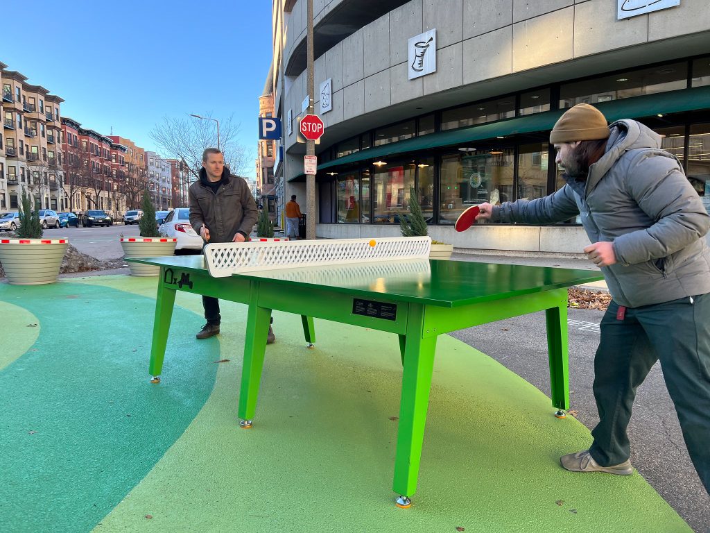 Two people play ping pong on a plaza closed to cars