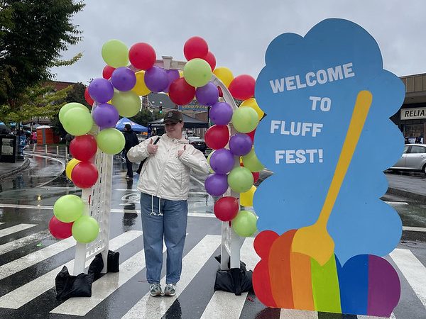 A person stands under a balloon covered archway next to a sign that reads "Welcome to Fluff Fest"