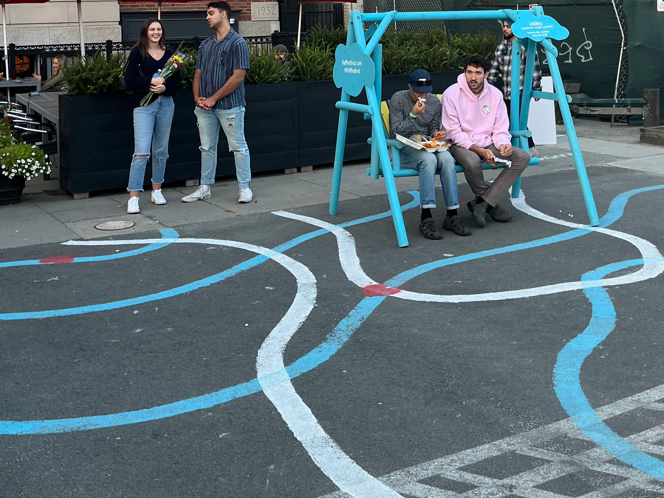 a plaza with white and blue painted lines on the ground. Two people sit on a blue swing.
