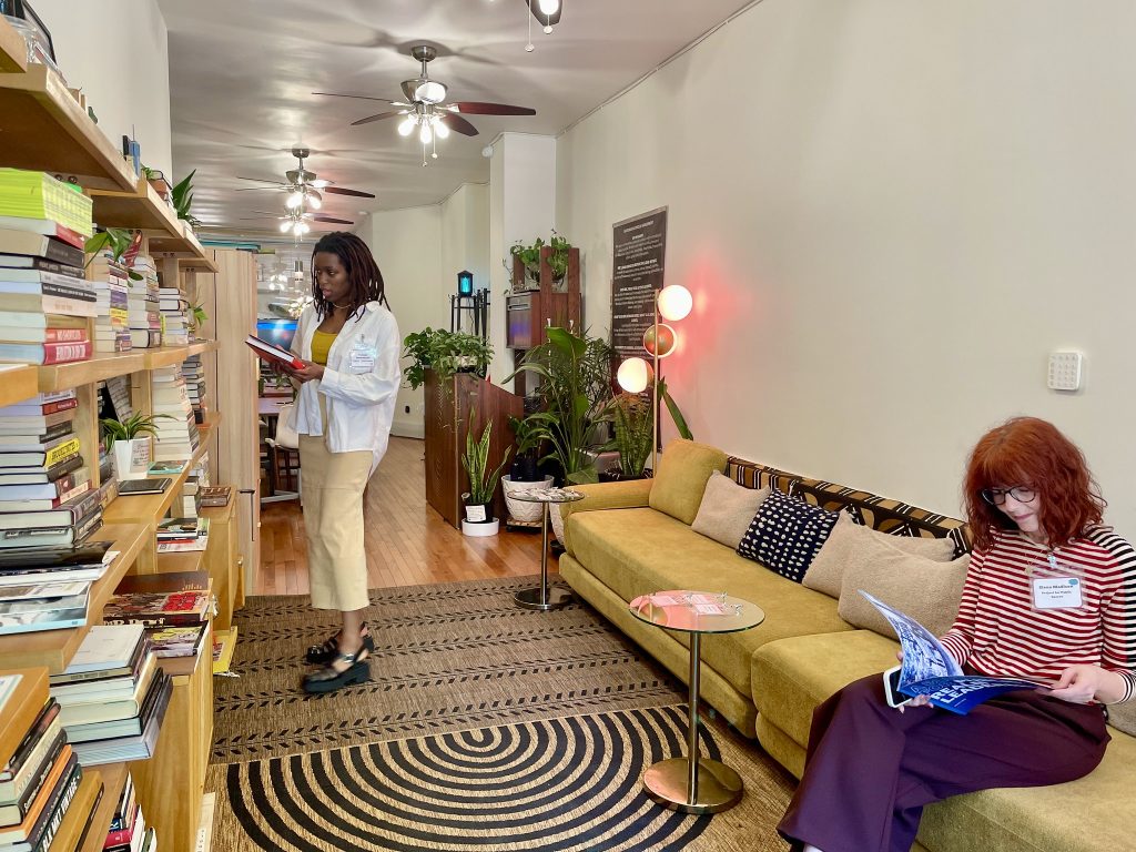 A person standing reading a book and another person sitting on a couch in a warm community space.